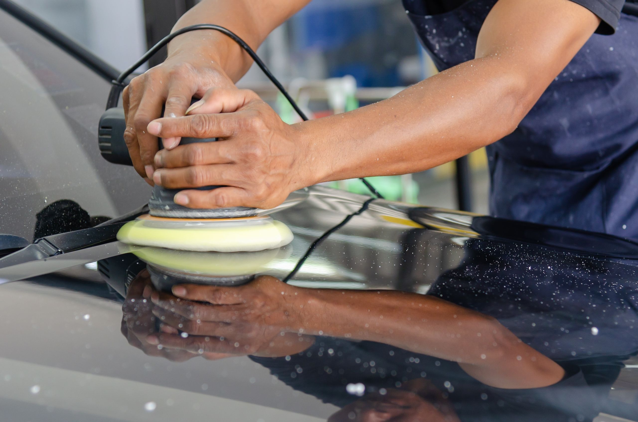 Detailer guiding a dual-action polisher with a yellow foam pad across a dark car hood, polish spatter flecked on the paint