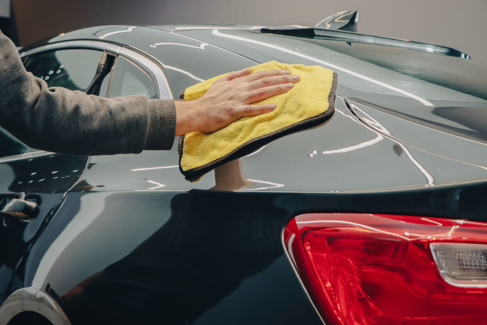 Hand buffing a yellow microfiber cloth across the glossy paint of a dark car, the by-hand application a coat of wax requires