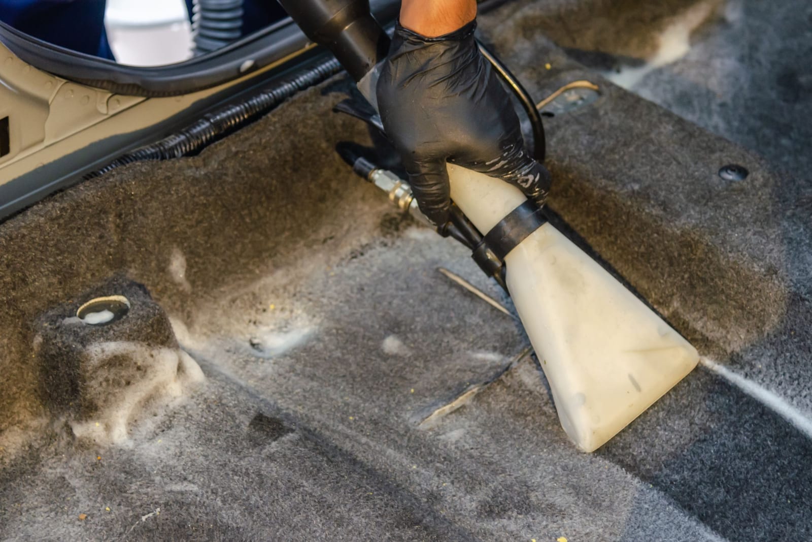 Gloved hand working an extraction wand across a car's dirty floor carpet, pulling embedded grit out of the pile