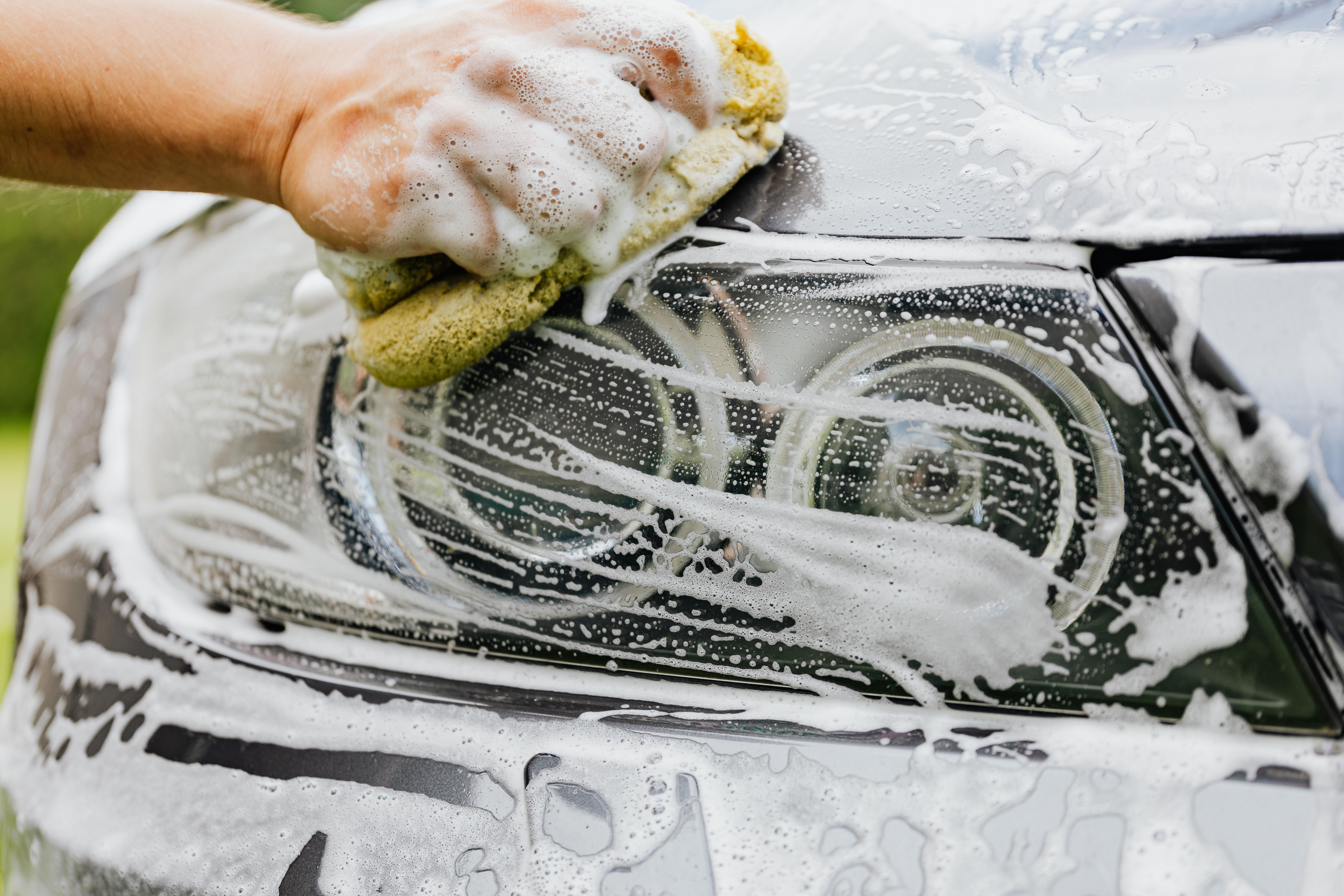 Bare hand pressing a foam-loaded yellow sponge across a car's headlight and front bumper, thick white lather streaming down the paint during a hand wash